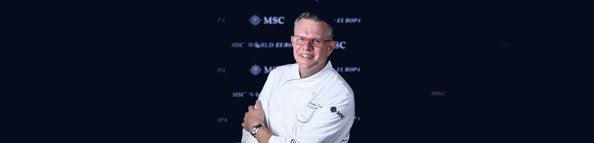 A chef in an MSC-branded uniform posing against a dark background with MSC logos | MSC Cruises A chef in an MSC-branded uniform posing against a dark background with MSC logos | MSC Cruises